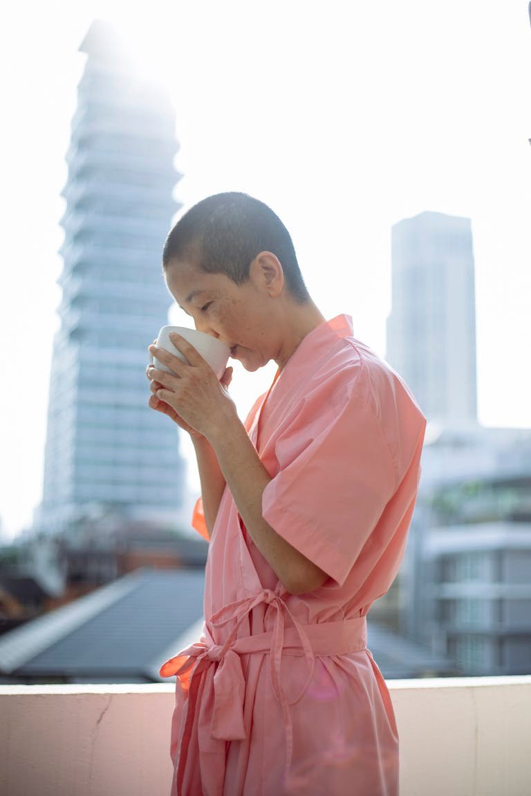 A woman in a pink robe enjoys a warm drink on a sunny day with tall buildings in the background.